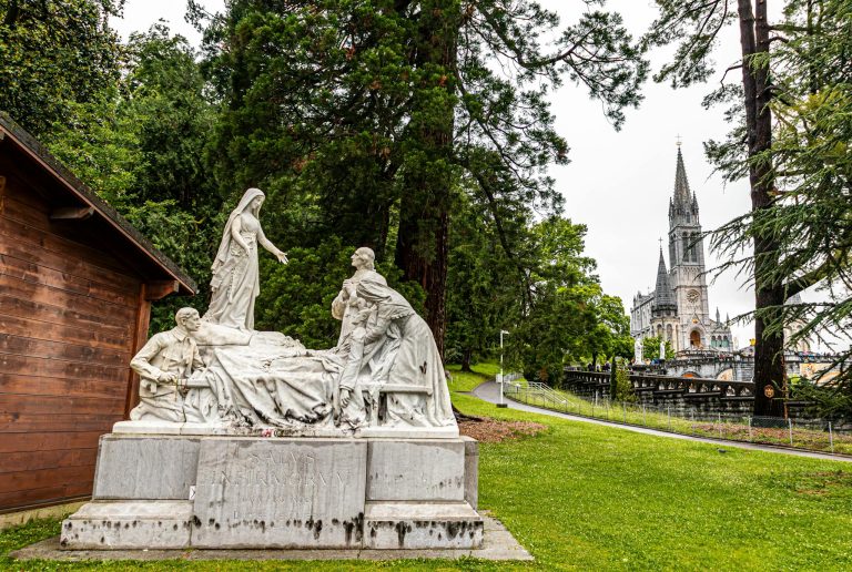 Marble statue with the Basilica of Our Lady of Lourdes in the background, amidst lush greenery.